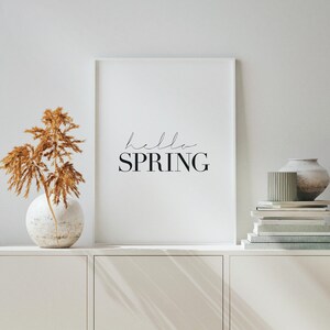 May include: A white framed print with the text "hello spring" in black lettering. The print is hanging on a white wall above a white cabinet. A vase with dried brown pampas grass is on the cabinet.