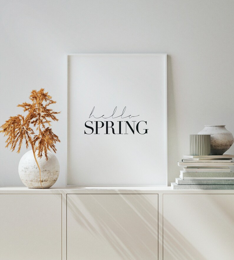 May include: A white framed print with the text "hello spring" in black lettering. The print is hanging on a white wall above a white cabinet. A vase with dried brown pampas grass is on the cabinet.