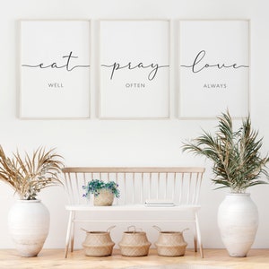 May include: Three framed prints with white backgrounds and black text. The prints read "eat well", "pray often", and "love always". The prints are hanging on a white wall above a white bench with a wooden seat.