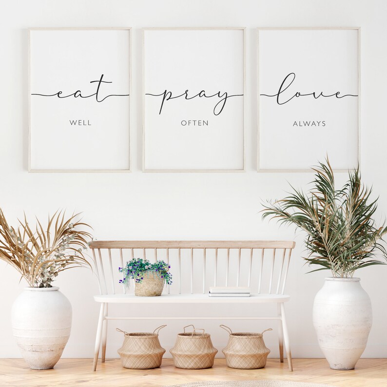 May include: Three framed prints with white backgrounds and black text. The prints read "eat well", "pray often", and "love always". The prints are hanging on a white wall above a white bench with a wooden seat.