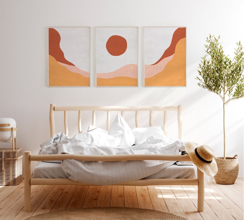 May include: Three abstract landscape prints in a minimalist style with a white background, orange and red hills, and a red circle in the center print. The prints are hanging above a wooden bed frame with white bedding.