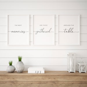 May include: Three framed prints with white frames and black text that reads "THE BEST memories", "ARE MADE gathered", and "AROUND THE table". The prints are displayed above a wooden table with decorative vases, books, and lanterns.