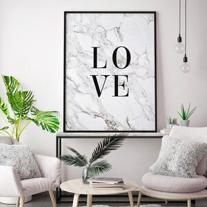 May include: A black framed print with a white marble background and the word "LOVE" in black text. The print is hanging on a white wall above a black console table with two pink chairs and a round wooden coffee table.