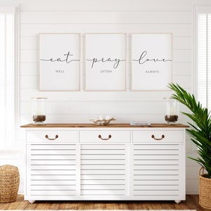May include: Three framed prints with white backgrounds and black calligraphy. The prints read "eat well", "pray often", and "love always". The prints are hanging on a white shiplap wall above a white dresser with three drawers.