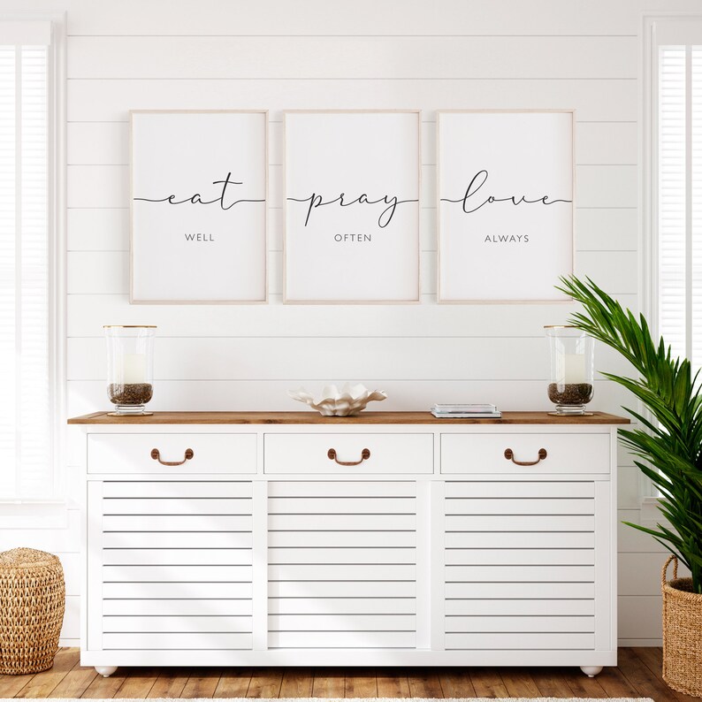 May include: Three framed prints with white backgrounds and black calligraphy. The prints read "eat well", "pray often", and "love always". The prints are hanging on a white shiplap wall above a white dresser with three drawers.
