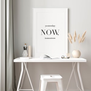 Peut inclure: Une impression encadr&eacute;e blanche avec le texte "yesterday-NOW. tomorrow" accroch&eacute;e &agrave; un mur blanc au-dessus d'un bureau blanc avec un tabouret blanc. Le bureau a un livre, des lunettes et un vase avec de l'herbe de pampa s&eacute;ch&eacute;e dessus.