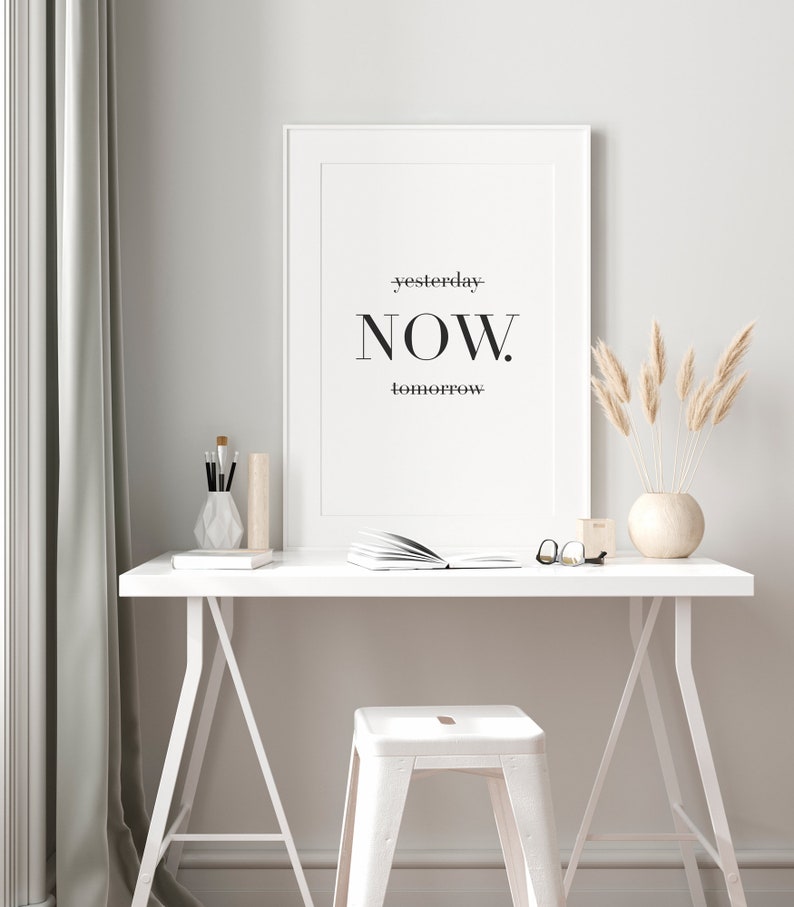 Peut inclure: Une impression encadr&eacute;e blanche avec le texte "yesterday-NOW. tomorrow" accroch&eacute;e &agrave; un mur blanc au-dessus d'un bureau blanc avec un tabouret blanc. Le bureau a un livre, des lunettes et un vase avec de l'herbe de pampa s&eacute;ch&eacute;e dessus.