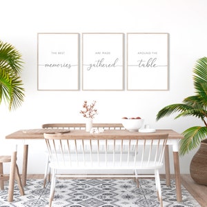 May include: Three framed prints with the text "THE BEST memories", "ARE MADE gathered", and "AROUND THE table" in a stylish script. The prints are displayed above a wooden dining table with white chairs, creating a cosy dining room setting.