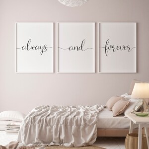 May include: Three white framed prints with black calligraphy on a light pink wall. The prints read "always", "and", and "forever". The prints are hanging above a bed with a white linen duvet and a light brown rug.