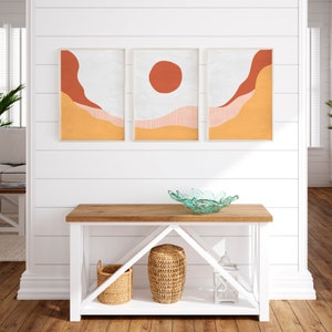 May include: A white console table with two shelves, a light wood top, and a white background. The table has a bowl on the top shelf and two baskets and a lantern on the bottom shelf. Three framed prints with abstract geometric shapes in orange, red, and white are hanging on the wall above the table.