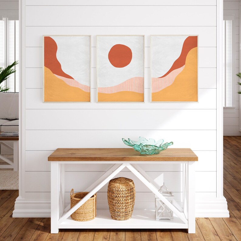 May include: A white console table with two shelves, a light wood top, and a white background. The table has a bowl on the top shelf and two baskets and a lantern on the bottom shelf. Three framed prints with abstract geometric shapes in orange, red, and white are hanging on the wall above the table.