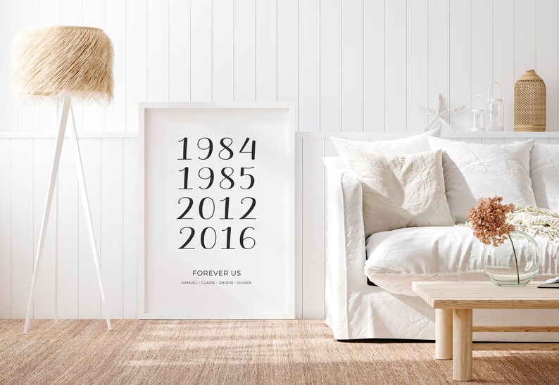 May include: A white framed print with black text that reads "1984, 1985, 2012, 2016, FOREVER US, SAMUEL, CLAIRE, SHORE, OLIVER" hanging on a white wall in a living room setting.