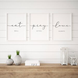 May include: Three white framed prints with black calligraphy on a white shiplap wall. The prints read "eat well", "pray often", and "love always". The prints are displayed above a wooden shelf with two white ceramic vases, a stack of books, and two white lanterns.