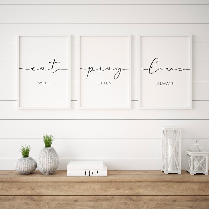 May include: Three white framed prints with black calligraphy on a white shiplap wall. The prints read "eat well", "pray often", and "love always". The prints are displayed above a wooden shelf with two white ceramic vases, a stack of books, and two white lanterns.