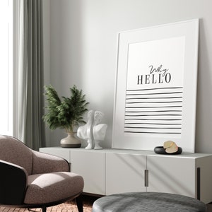 May include: A white framed print with black text that reads "Why HELLO" and black horizontal lines on a white background. The print is hanging on a white wall above a white cabinet with two doors. There are two white statues and a green plant in a brown vase on the cabinet.