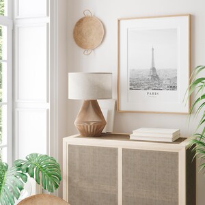 Peut inclure: Une lampe blanche avec un abat-jour beige est pos&eacute;e sur un meuble en bois avec une fa&ccedil;ade en rotin tiss&eacute;. Une photographie en noir et blanc encadr&eacute;e de la tour Eiffel avec le mot "PARIS" imprim&eacute; en dessous est accroch&eacute;e au mur au-dessus du meuble. Un panier tiss&eacute; est accroch&eacute; au mur &agrave; gauche de la photographie.