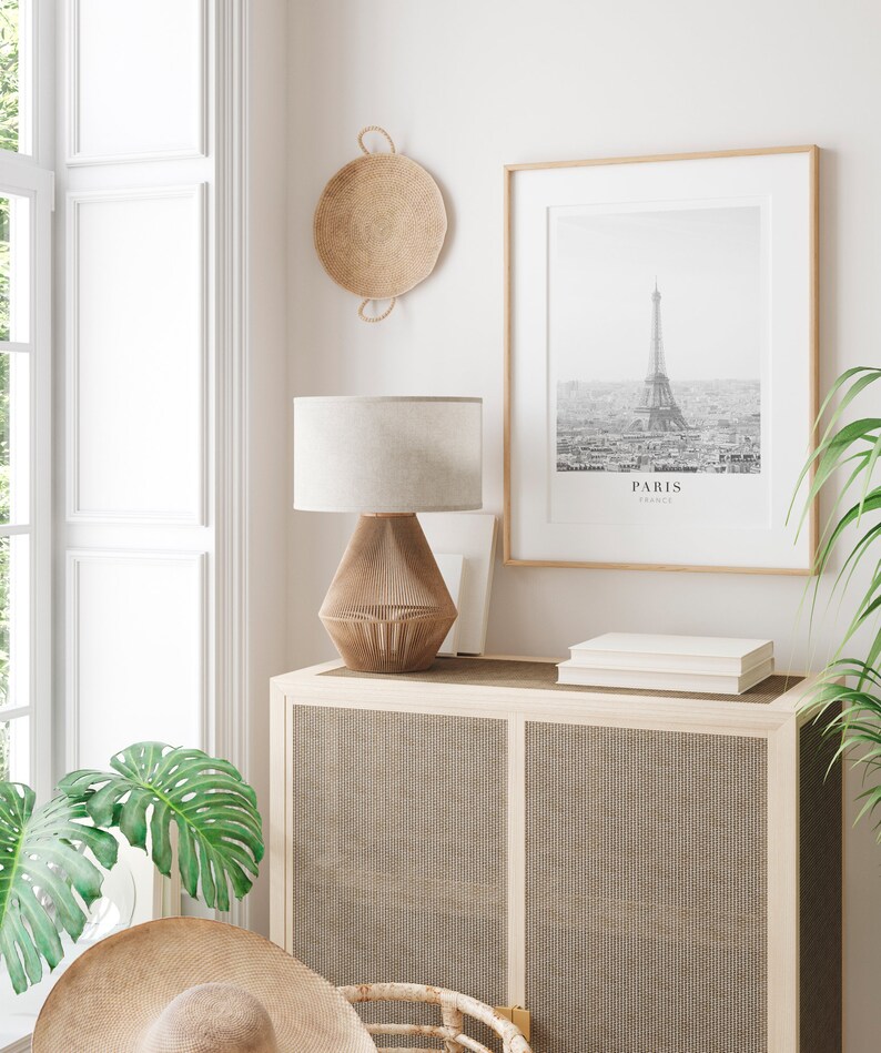 Peut inclure: Une lampe blanche avec un abat-jour beige est pos&eacute;e sur un meuble en bois avec une fa&ccedil;ade en rotin tiss&eacute;. Une photographie en noir et blanc encadr&eacute;e de la tour Eiffel avec le mot "PARIS" imprim&eacute; en dessous est accroch&eacute;e au mur au-dessus du meuble. Un panier tiss&eacute; est accroch&eacute; au mur &agrave; gauche de la photographie.