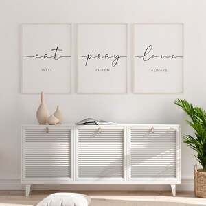 May include: Three framed prints with white mats and black calligraphy. The prints read "eat well", "pray often", and "love always". The prints are hanging on a white wall above a white cabinet with three doors.