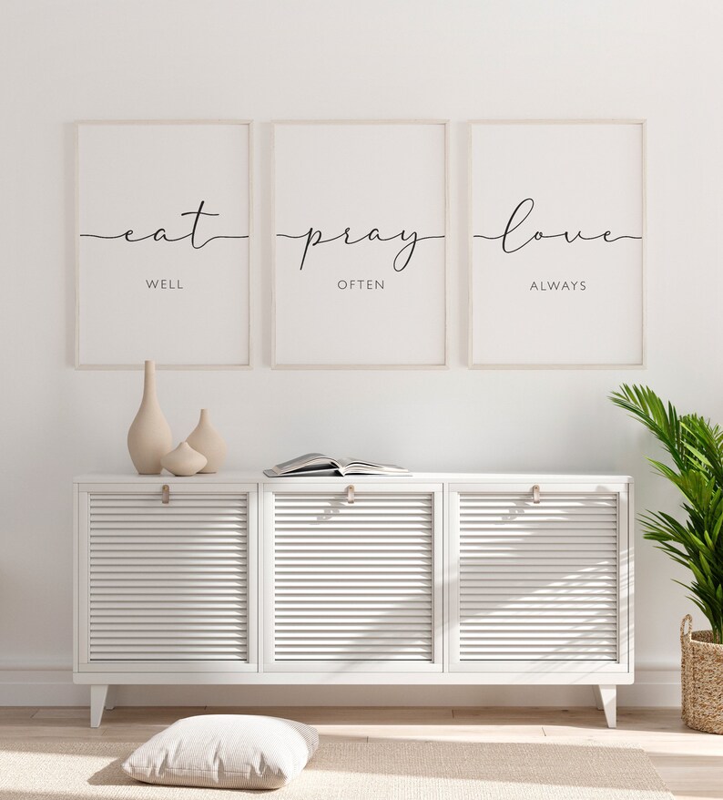 May include: Three framed prints with white mats and black calligraphy. The prints read "eat well", "pray often", and "love always". The prints are hanging on a white wall above a white cabinet with three doors.