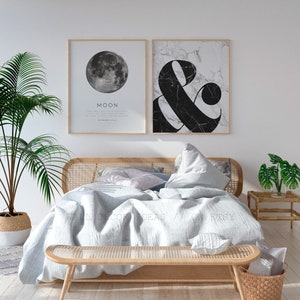 Op de afbeelding: Twee ingelijste prints op een witte muur boven een bed met een lichtgrijs linnen dekbed en een geweven houten hoofdeinde. De print aan de linkerkant toont een zwart-witte afbeelding van de maan met het woord "MAAN" en een citaat eronder. De print aan de rechterkant toont een zwart-witte afbeelding van een ampersand-symbool op een marmeren achtergrond.