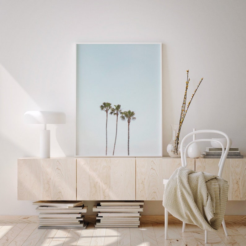 May include: A framed print of three palm trees against a light blue sky, hanging above a light wood cabinet with stacked books and a white chair with a white throw blanket.