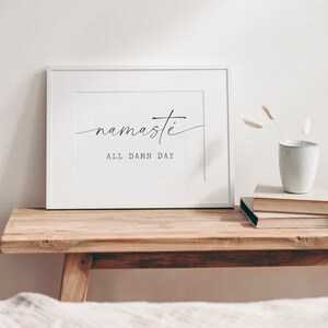 K&ouml;nnte beinhalten: Wei&szlig;er gerahmter Druck mit dem Text "namaste all damn day" in schwarzer Kalligraphie auf wei&szlig;em Hintergrund. Der Druck liegt auf einem Holztisch mit einer Vase mit Trockenblumen und einem Stapel B&uuml;cher.