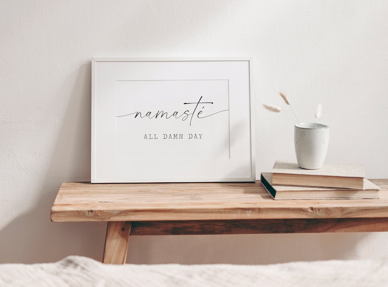 K&ouml;nnte beinhalten: Wei&szlig;er gerahmter Druck mit dem Text "namaste all damn day" in schwarzer Kalligraphie auf wei&szlig;em Hintergrund. Der Druck liegt auf einem Holztisch mit einer Vase mit Trockenblumen und einem Stapel B&uuml;cher.