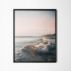 May include: A close-up of a wave breaking on a beach, with the sun setting in the background. The water is a light blue colour, and the sand is a light brown colour.