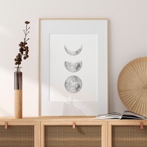 May include: A framed print of three watercolor moon phases in shades of gray on a white background. The print is hanging on a wall above a wooden cabinet with a woven rattan front.