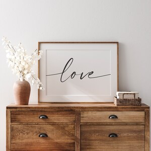 Peut inclure: Une impression encadr&eacute;e avec le mot "love" &eacute;crit en cursive sur un fond blanc. L'impression est accroch&eacute;e &agrave; un mur blanc au-dessus d'une commode en bois &agrave; deux tiroirs.