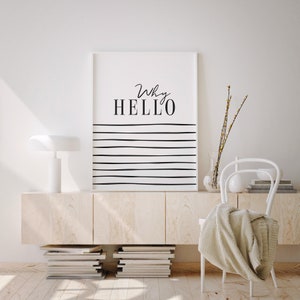 May include: A white framed print with black text that reads "Why HELLO" and black horizontal lines on a white background. The print is hanging on a white wall above a light wood cabinet with a white lamp on top. A white chair with a beige throw blanket is in the foreground.