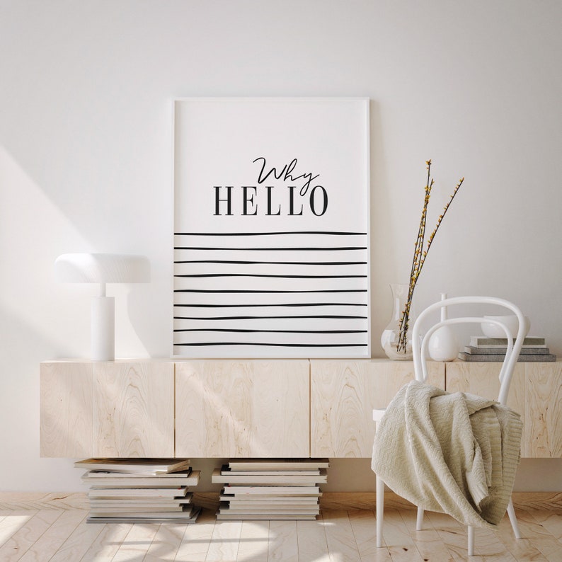 May include: A white framed print with black text that reads "Why HELLO" and black horizontal lines on a white background. The print is hanging on a white wall above a light wood cabinet with a white lamp on top. A white chair with a beige throw blanket is in the foreground.