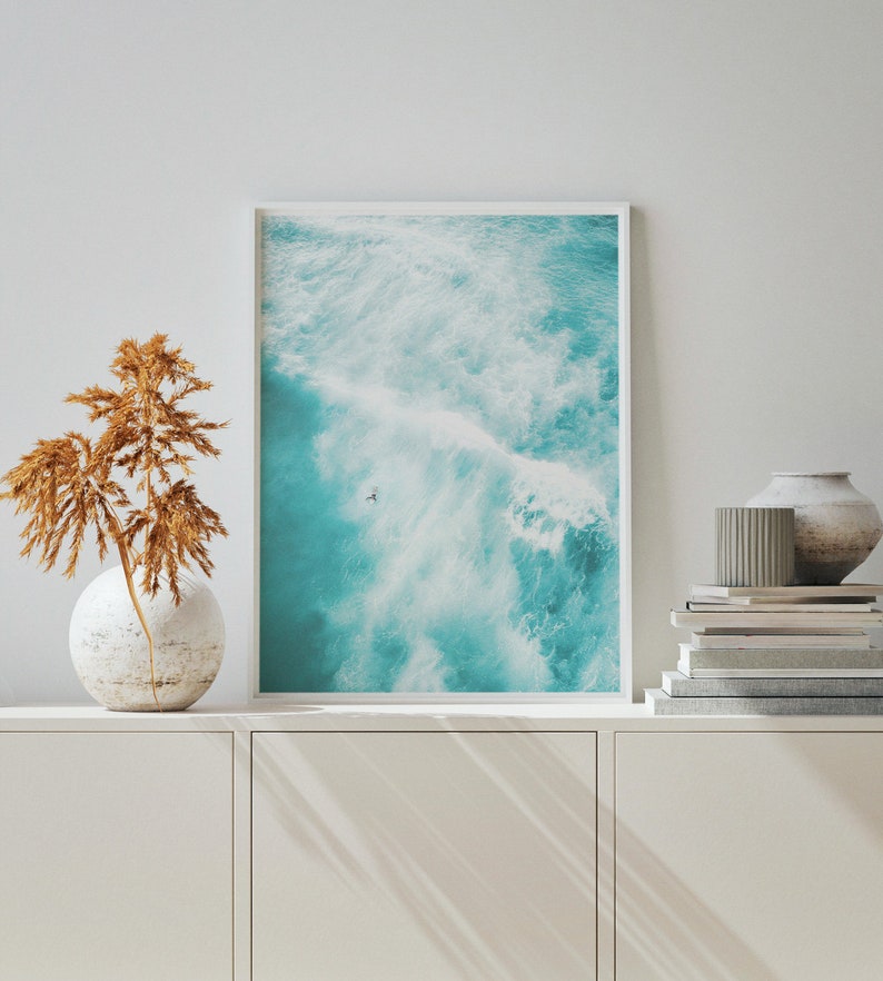 May include: A framed photograph of a turquoise ocean with white foamy waves. The photo is hanging on a white wall above a white cabinet.