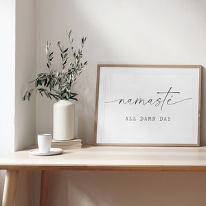 K&ouml;nnte beinhalten: Ein gerahmter Druck mit dem Text "namaste all damn day" in schwarzer Schrift auf wei&szlig;em Hintergrund. Der Druck h&auml;ngt an einem hellen Holzregal an der Wand &uuml;ber einem Holztisch mit einer wei&szlig;en Vase und einer gr&uuml;nen Pflanze.