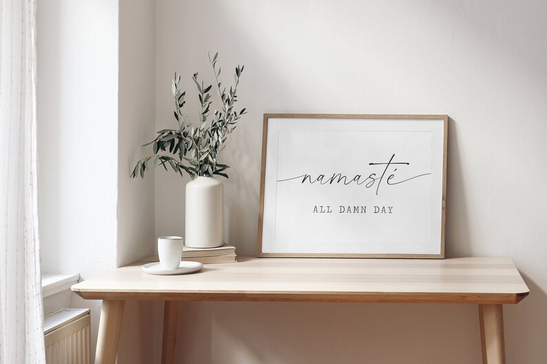 K&ouml;nnte beinhalten: Ein gerahmter Druck mit dem Text "namaste all damn day" in schwarzer Schrift auf wei&szlig;em Hintergrund. Der Druck h&auml;ngt an einem hellen Holzregal an der Wand &uuml;ber einem Holztisch mit einer wei&szlig;en Vase und einer gr&uuml;nen Pflanze.