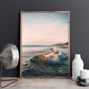 May include: A framed photograph of a wave breaking on a beach at sunset. The water is a deep blue and the sky is a soft pink and orange.