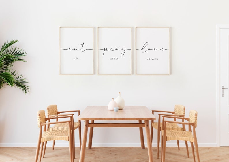 May include: Three framed prints with the text "eat well, pray often, love always" hanging on a white wall above a wooden table with two chairs.