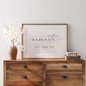 K&ouml;nnte beinhalten: Ein gerahmter Druck mit dem Text "namaste all damn day" in einer minimalistischen schwarzen Schrift auf wei&szlig;em Hintergrund. Der Druck h&auml;ngt an einer wei&szlig;en Wand &uuml;ber einer Kommode aus Holz.