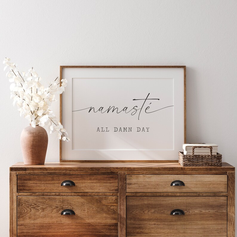K&ouml;nnte beinhalten: Ein gerahmter Druck mit dem Text "namaste all damn day" in einer minimalistischen schwarzen Schrift auf wei&szlig;em Hintergrund. Der Druck h&auml;ngt an einer wei&szlig;en Wand &uuml;ber einer Kommode aus Holz.