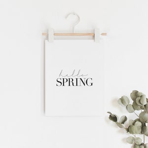 May include: A white framed print with the text "hello spring" in black script font. The print is hanging on a white wall with a wooden dowel and a white hook. A sprig of eucalyptus leaves is in the bottom right corner of the image.