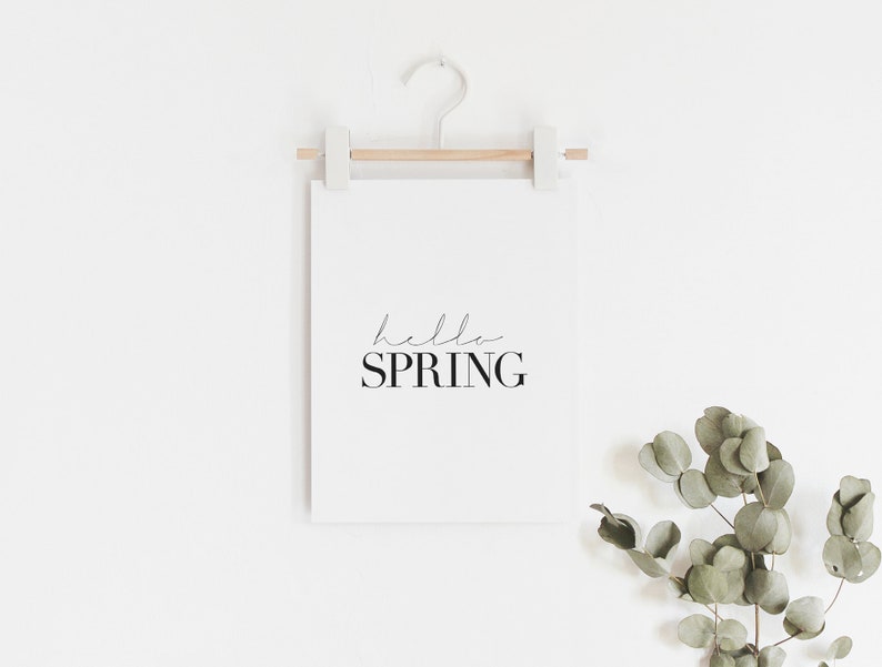 May include: A white framed print with the text "hello spring" in black script font. The print is hanging on a white wall with a wooden dowel and a white hook. A sprig of eucalyptus leaves is in the bottom right corner of the image.