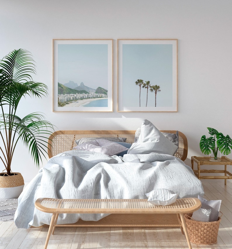 May include: A white bed with a light blue linen duvet and a woven headboard. The bed is styled with pillows and a throw blanket. Two framed prints of beach scenes hang above the bed. A woven bench sits at the foot of the bed.