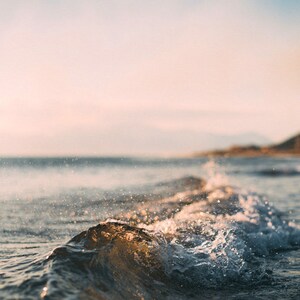 May include: A close-up of a wave breaking on a sandy beach. The water is a light blue and the sand is a light brown. The sky is a light blue with clouds in the distance.
