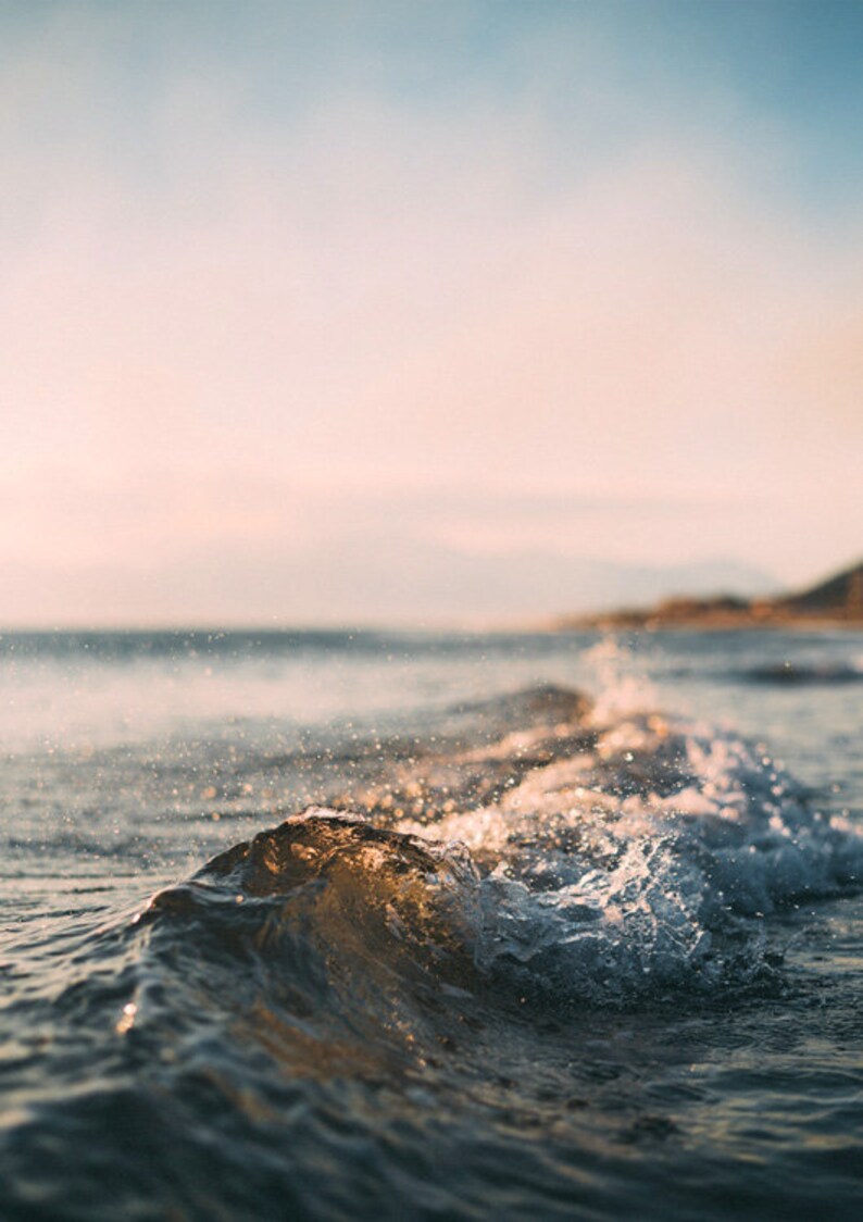 May include: A close-up of a wave breaking on a sandy beach. The water is a light blue and the sand is a light brown. The sky is a light blue with clouds in the distance.