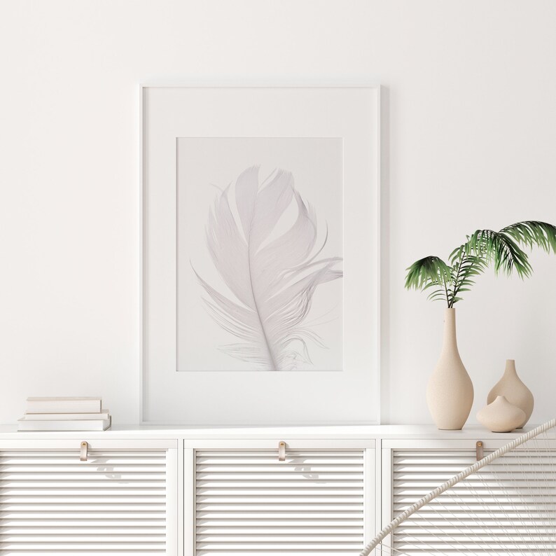 Peut inclure: Une impression encadr&eacute;e blanche d'une seule plume blanche sur fond blanc. L'impression est accroch&eacute;e &agrave; un mur blanc au-dessus d'une armoire blanche &agrave; deux portes. Il y a deux vases avec des plantes vertes sur l'armoire.