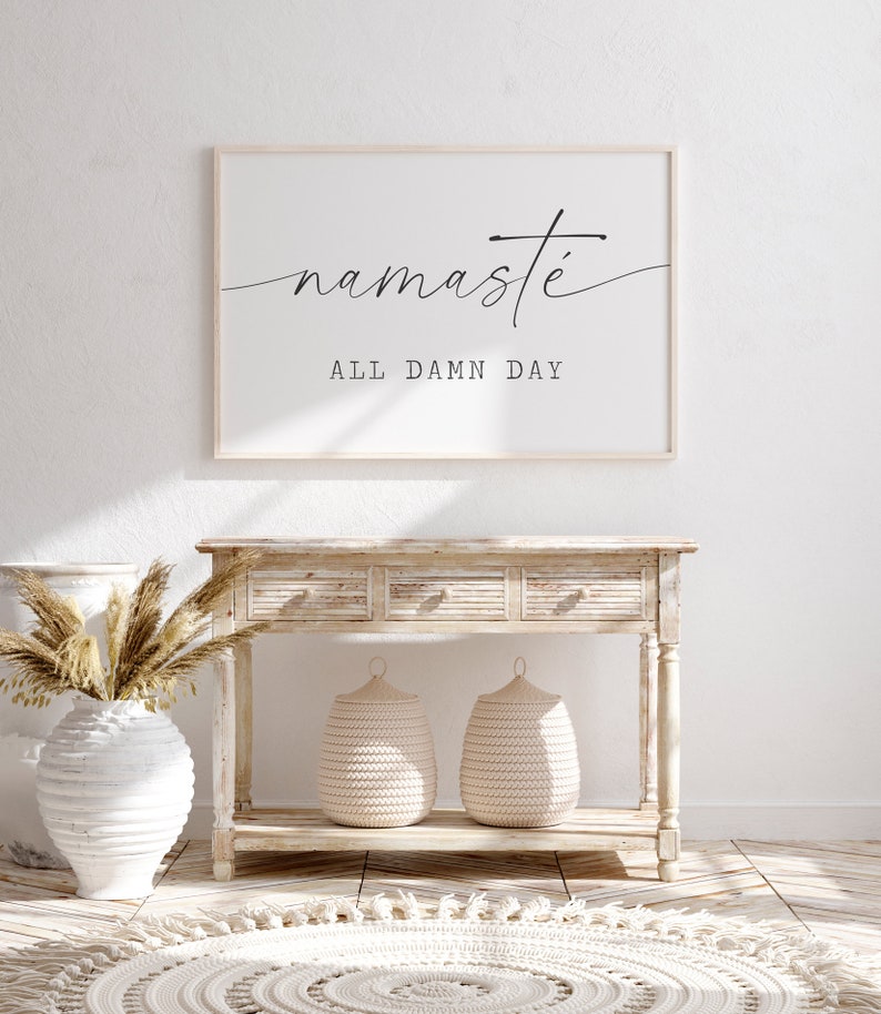 K&ouml;nnte beinhalten: Ein wei&szlig;er Holz-Konsolentisch mit drei Schubladen und zwei geflochtenen K&ouml;rben auf der unteren Ablage. Ein gerahmtes Bild mit dem Text "namaste all damn day" h&auml;ngt an der Wand hinter dem Tisch. Eine wei&szlig;e Vase mit getrocknetem Pampasgras steht auf dem Boden vor dem Tisch.
