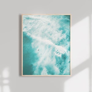 May include: An aerial view of a turquoise ocean with white waves crashing. A lone figure is seen surfing in the distance.