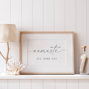 K&ouml;nnte beinhalten: Ein gerahmter Druck mit dem Text "namaste all damn day" in einer minimalistischen schwarzen Schrift auf wei&szlig;em Hintergrund. Der Druck wird auf einem wei&szlig;en Regal mit einer Holzlampe und einer Glasflasche pr&auml;sentiert.