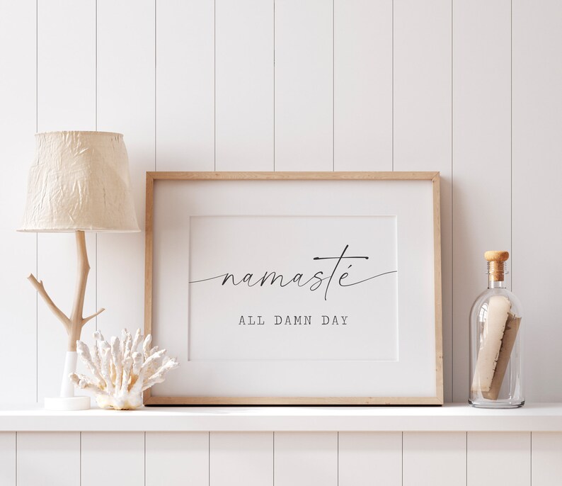 K&ouml;nnte beinhalten: Ein gerahmter Druck mit dem Text "namaste all damn day" in einer minimalistischen schwarzen Schrift auf wei&szlig;em Hintergrund. Der Druck wird auf einem wei&szlig;en Regal mit einer Holzlampe und einer Glasflasche pr&auml;sentiert.