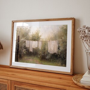 May include: A framed painting of a rural landscape with white clothes hanging on a line to dry. The painting is in a wooden frame and has a soft, muted color palette.